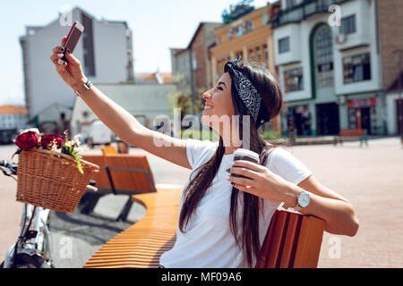 Lächelnden jungen Frau unter selfie, sitzen auf einer Bank an einem sonnigen Tag, neben dem Fahrrad mit Blumenkorb. Stockfoto