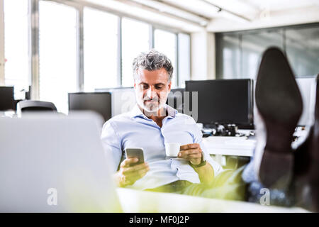 Entspannt Reife Unternehmer sitzen am Schreibtisch im Büro mit Handy und trinken Kaffee Stockfoto