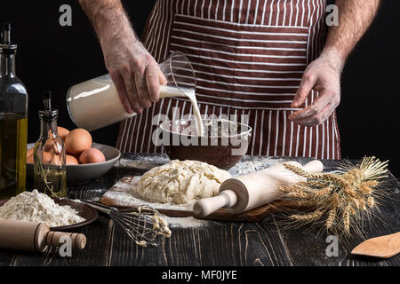 Baker in Uniform gießen Milch aus Kunststoff Flasche in die Schüssel. Eine Handvoll Mehl mit Ei auf eine rustikale Küche. Stockfoto