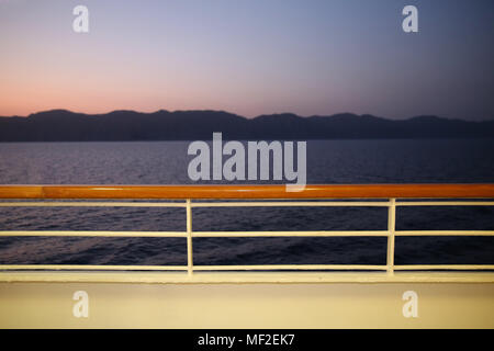 Blick vom Deck eines Kreuzfahrtschiffes in der Dämmerung in Richtung Küste. Es ist pink und lila Farben in den Himmel über einem ruhigen Meer. Stockfoto