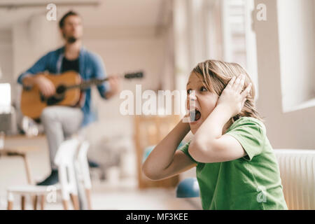 Sohn entsetzt über seine Ohren mit Vater Gitarre spielen zu Hause Stockfoto