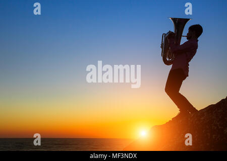 Silhouette des Musikers mit der Tuba/Trompete auf felsigen Küste bei Sonnenuntergang. Stockfoto