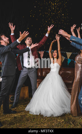 Fröhliche Hochzeit paar Freunden und heben ihre Arme während Konfetti fällt über ihre Köpfe in der Nacht Party im Freien Stockfoto
