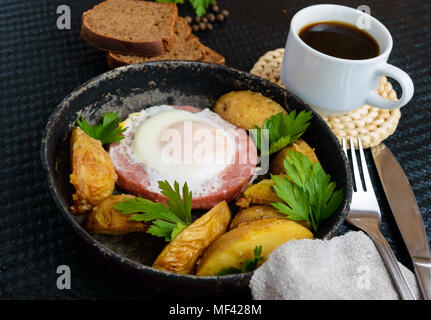 Spiegelei in Salami und gebackene Kartoffeln. Einreichung von eine Pfanne mit einer Tasse Kaffee und Roggenbrot. Frühstück. Stockfoto