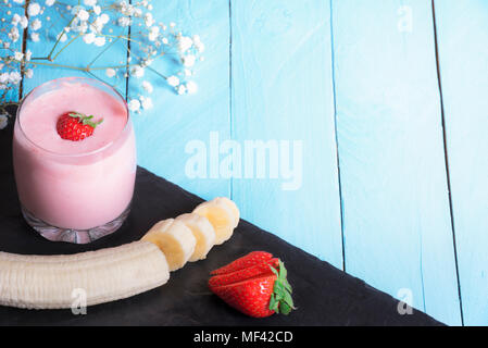 Gesundes Konzept Essen mit ein Glas frische Erdbeeren und Bananen Smoothie, geschnittene Früchte auf einer schwarzen Platte, auf einem blauen Hintergrund, im Sonnenlicht. Stockfoto