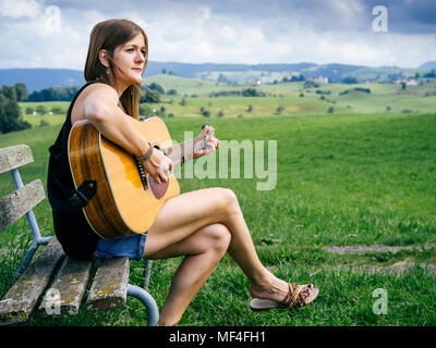 Foto einer jungen Frau spielen akustische Gitarre auf einer Parkbank. Stockfoto