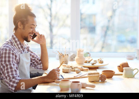 Porträt der jungen gemischten Rennen männlichen Keramiker sprechen auf Smartphone und fröhlich lächelnd beim Sitzen am Tisch mit Ton Kunsthandwerk Stockfoto