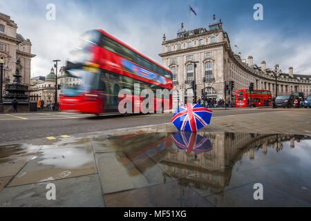 London, England - 03.15.2018: Reflexion der roten Doppeldecker Busse unterwegs am Piccadilly Circus mit britischen Regenschirm. Piccadilly Circus ist der m Stockfoto