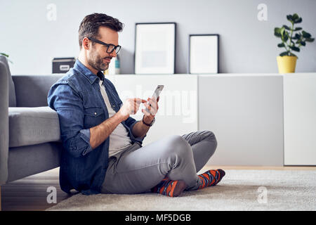 Lächelnd Mann sitzt auf dem Fußboden im Wohnzimmer mit Handy Zimmer Stockfoto