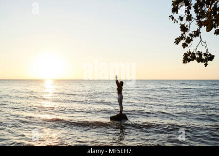 Junge Frau mit Yoga auf einem Felsen im Meer bei Sonnenuntergang Stockfoto