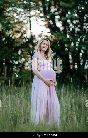 Eine schwangere Frau, die in einem Sommer Kleid ihr Bauch berühren. Ein Porträt eines lächelnden erwartet Mutter ihre Schwangerschaft bump streicheln. Stockfoto