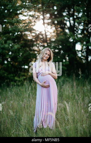 Eine Vorderansicht eines glücklichen schwangeren Frau in einem Sommer Kleid ihr Bauch berühren. Ein Porträt eines lächelnden erwartet Mutter ihre Schwangerschaft bump streicheln. Stockfoto