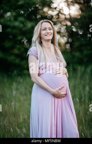 Eine junge werdende Mutter sanft berühren ihre Schwangerschaft Bauch. Ein Porträt eines lächelnden Schwangere genießen in der Natur an einem Sommerabend. Stockfoto