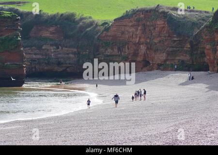 Urlauber auf einem Kieselstrand mit roten Otter Sandstein Geologie der Jurassic Coast. Ladram Bay Holiday Park, East Devon, Großbritannien. April, 2018. Stockfoto