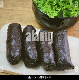 Japanische Küche, Traditionelle ungeschnitten Japanischen Reis Maki Sushi Roll mit Nori Algen auf Schneidebrett serviert mit grünen Eichen. Stockfoto