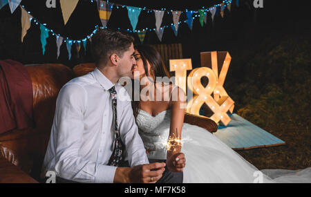 Romantische Hochzeit paar sitzt auf einem Sofa zu küssen, während sie Wunderkerzen in der Hand auf einem Night Party im Freien Stockfoto