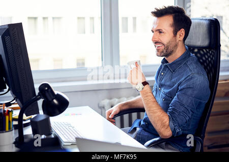 Lächelnd Mann am Computer und Kaffee trinken am Schreibtisch im Büro suchen Stockfoto