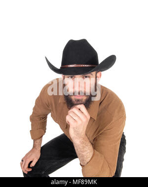 Ein junger Mann sitzt auf einem Stuhl bücken nach vorne trägt einen schwarzen Cowboyhut und mit der anderen Hand am Kinn, auf weißem Hintergrund Stockfoto