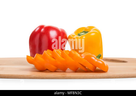 Eine orange Paprika, in Scheiben geschnitten, sitzt auf einem Bambus Schneidebrett. Andere farbige Paprika sind in der Nähe. Stockfoto