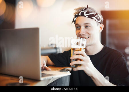 Junge lächelnde Mann hält ein Glas Bier und Arbeiten am Laptop. Stockfoto
