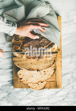 Weibliche Hände schneiden frisch gebackenem Sauerteigbrot, vertikale Zusammensetzung Stockfoto