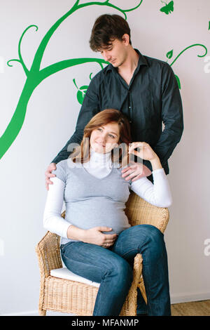 Schwangerschaft, Familie, Behandlungskonzept. Groß und gut aussehender Mann mit dunklen Haare hinter seine schwangere Frau, die sich in den Korbstuhl wi Sitzt steht. Stockfoto