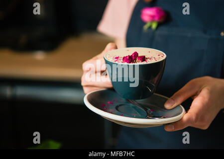 Tasse und Untertasse mit Kaffee, Schaum, Milch und Pink Flower Buds. Tasse raf rose Kaffee Barista Hände. Stockfoto