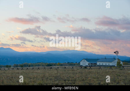 Ein Mountain Ranch, komplett mit Pferdestall und Windmühle hinter einem Zaun, Uhren die Sonne hinter dem Moskitonetz Berge. Stockfoto