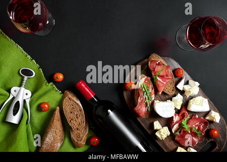 Schinken, Salami, Baguette, Scheiben, Tomaten und die Mutternder rustikalen Holzbrett, zwei Gläser Rotwein auf schwarzem Hintergrund Stockfoto