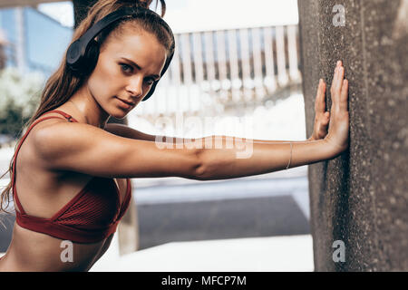 Porträt der jungen Frau das Tragen von Kopfhörern tun Training durch eine Wand im Freien. Weibliche Athleten tun warmup Übungen. Stockfoto