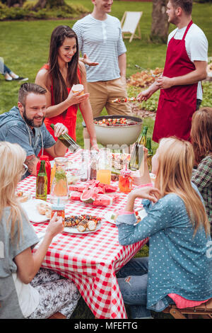 Eine Gruppe von Freunden im Garten genießen Sie eine Mahlzeit, Essen und Trinken durch eine Tabelle und Männer in der Zurück mehr shashliks auf dem Grill Stockfoto