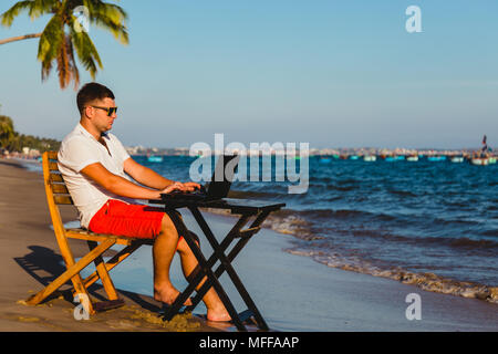 Mann bei der Arbeit mit einem Laptop, auf einer Hängematte am Strand. Konzept der digitale Nomade, externe Mitarbeiter, unabhängige Position Unternehmer. Stockfoto