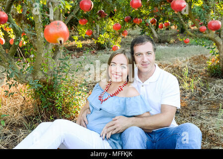 Portrait von Happy Family Rest in der pomegrate Obst Garten. Mann die Hand auf Magen, der seine schwangere Frau. Fruchtbarkeit Konzept. Sonnenuntergang li Stockfoto