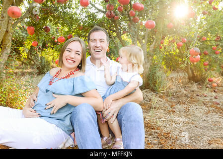 Portrait von Happy husbent und schwangere Frau mit niedlichen Baby Mädchen Tochter in Ruhe und Spaß in der pomegrate Obst Garten. Familie Zeit verbringen. Sonnen Stockfoto