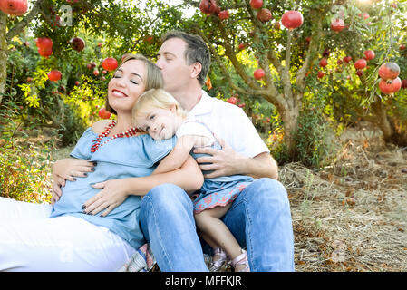Portrait von Happy husbent und schwangere Frau mit niedlichen Baby Mädchen Tochter in Ruhe und Spaß in der pomegrate Obst Garten. Familie Zeit verbringen. Sonnen Stockfoto