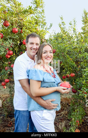 Portrait von Happy Family im pomegrate Garten. Mann, Hände auf Bauch von seiner schwangeren Frau mit reifen Früchten. Fruchtbarkeit Konzept. Sonnenuntergang lig Stockfoto