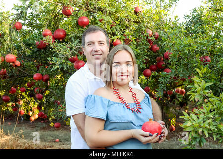Portrait von Happy Family im pomegrate Garten. Mann, Hände auf Bauch von seiner schwangeren Frau mit reifen Früchten. Fruchtbarkeit Konzept. Sonnenuntergang lig Stockfoto