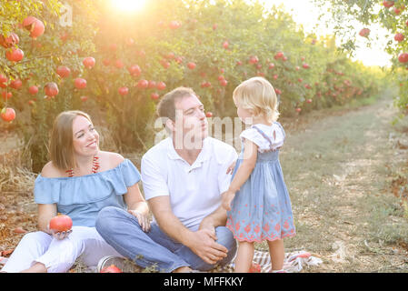 Portrait von lächelnden Eltern mit Baby Mädchen Tochter in Ruhe und Spaß in der pomegrate Obst Garten auf den Sonnenuntergang. Glückliche Familie Zeit verbringen. Sunse Stockfoto