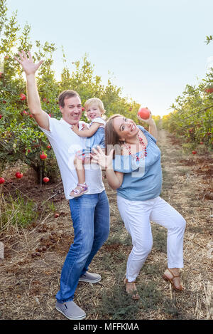 Portrait von lächelnden Eltern mit Baby Mädchen Tochter plaiying der Ape und Spaß in der pomegrate Obst Garten auf den Sonnenuntergang. Glückliche Familie Ausgaben Stockfoto