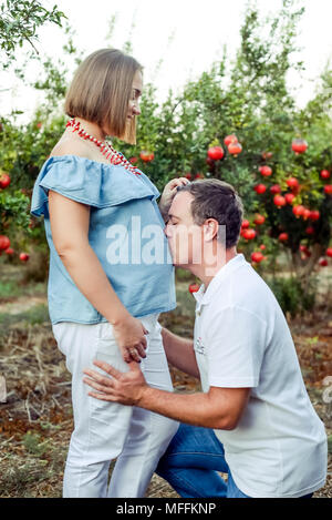 Vertikale Portrait von Happy Family im pomegrate Obst Garten. Mann kniend und küssen Magen von seiner schwangeren Frau. Fruchtbarkeit Konzept. Sonnenuntergang Stockfoto