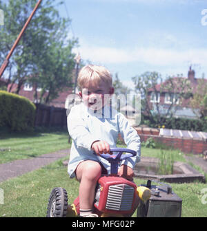 1960, historische, kleine blonde Junge, draußen im Garten gerne saß auf seinem trettraktor, England, UK. Stockfoto