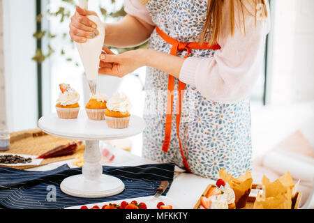 7/8-Ansicht von Konditor in melierter Schürze cupcakes Richtfest mit Sahne mit einem Spritzbeutel. Der cremige Kuchen Stockfoto