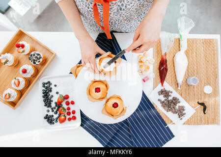 Blick von oben auf die 7/8-frau Hände verzieren berry Cupcakes mit Sahne auf dem Küchentisch gefüllt Stockfoto