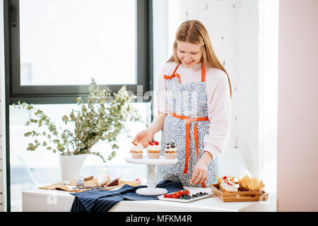 Schöne glückliche Frau angenehm lächelnd dekorieren Cupcakes mit Berry auf dem Tisch in gut beleuchteten Zimmer Stockfoto