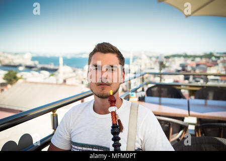 Ein junger stattlicher Mann mit Schnurrbart ist das Rauchen einer Wasserpfeife auf der Terrasse mit Blick auf die Stadt Stockfoto