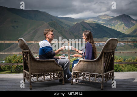 Junges Paar in gute Laune sitzt auf der Terrasse mit Tassen Kaffee am Morgen mit einem wunderschönen Blick auf die Berge. Hübsches Mädchen mit Freund in Pause Stockfoto