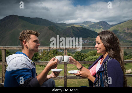 Junges Paar sitzt auf der Terrasse mit Tassen Kaffee am Morgen mit schöner Aussicht. Schöne Mädchen mit Freund in gute Stimmung beim Frühstück. Stockfoto