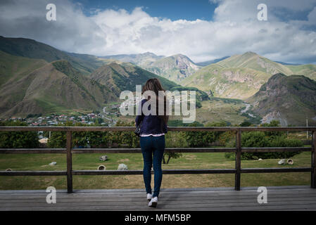 Junge Frau steht wieder auf die Aussichtsplattform mit herrlichem Blick auf die Berge und Ortschaft im Tal. Stockfoto