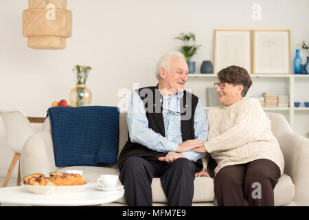 Gerne älteres Paar in Liebe, Zärtlichkeit und ihre Zeit zusammen halten sich an den Händen und Sitzen auf einem Sofa mit blauen Decke in ein gemütliches Wohnzimmer genießen r Stockfoto