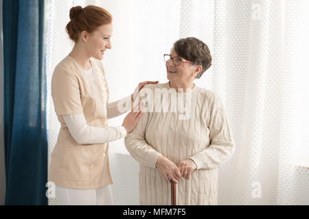 Professionelle Betreuer in Uniform und unterstützen eine ältere Frau mit einem Stock gegen weißen Hintergrund in einem Seniorenheim sprechen Stockfoto